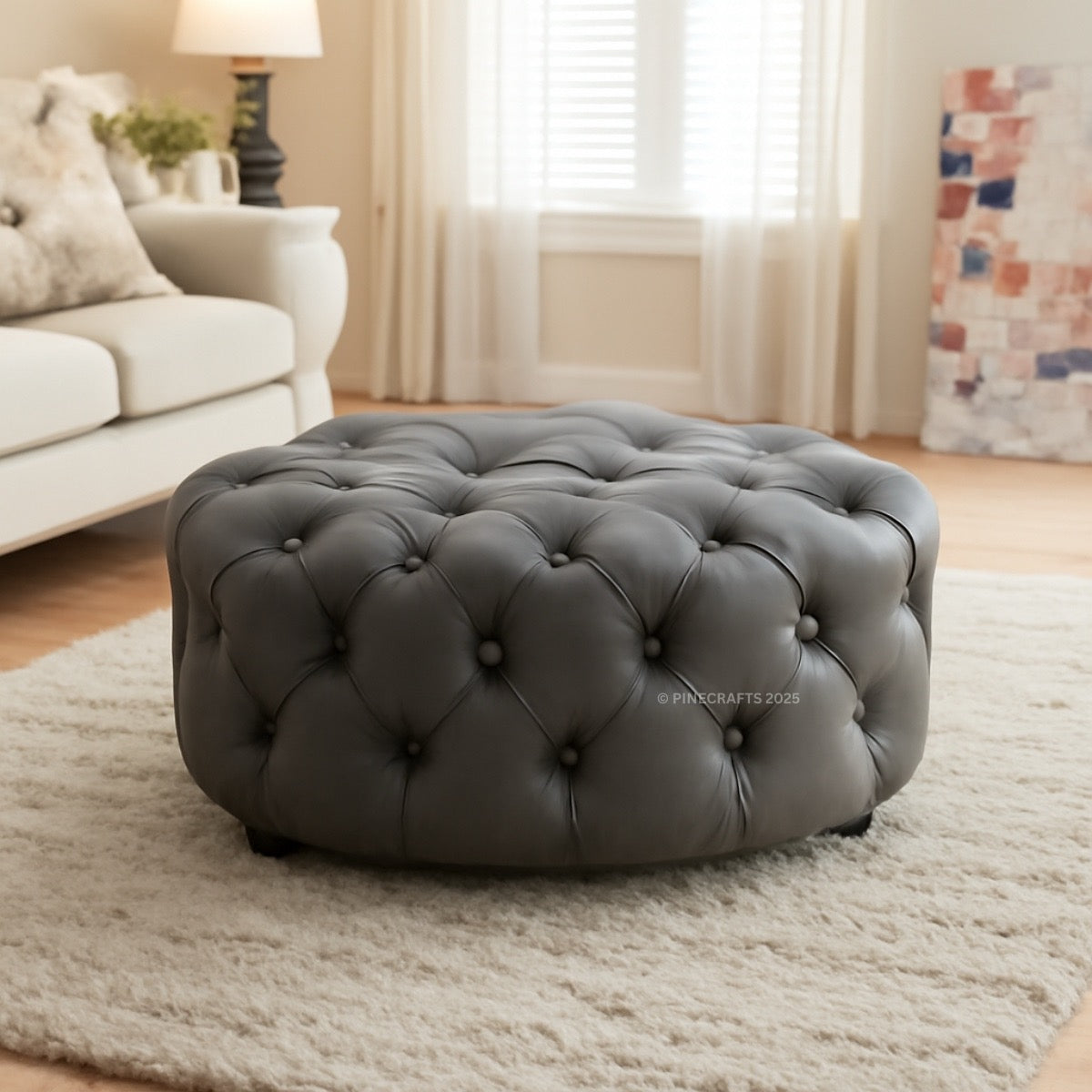 Gray tufted ottoman in a living room setting with a couch and lamp in the background.